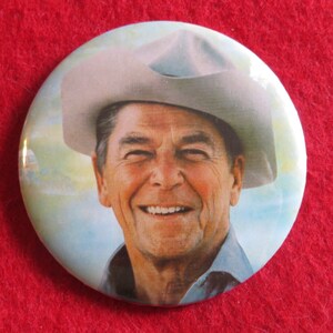 May include: A vintage button featuring a smiling man wearing a cowboy hat. The background is a light blue sky with clouds.