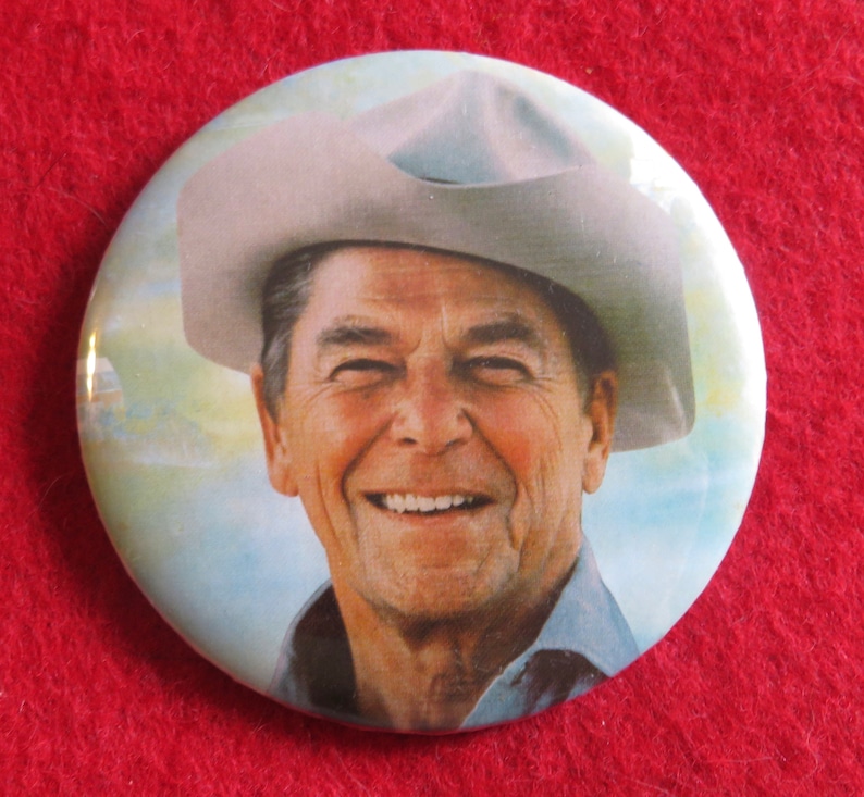 May include: A vintage button featuring a smiling man wearing a cowboy hat. The background is a light blue sky with clouds.