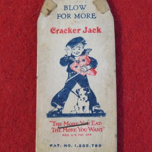 Może przedstawiać: Winietkowe pudełko Cracker Jack z niebiesko-białą ilustracją chłopca w marynarskim stroju trzymającego czerwone pudełko i psa. Na pudełku widnieje tekst "Blow for More Cracker Jack" oraz "The More You Eat-The More You Want" wraz z tekstem "Reg U.S. Pat. Off" i "Pat. No. 1,235,799".