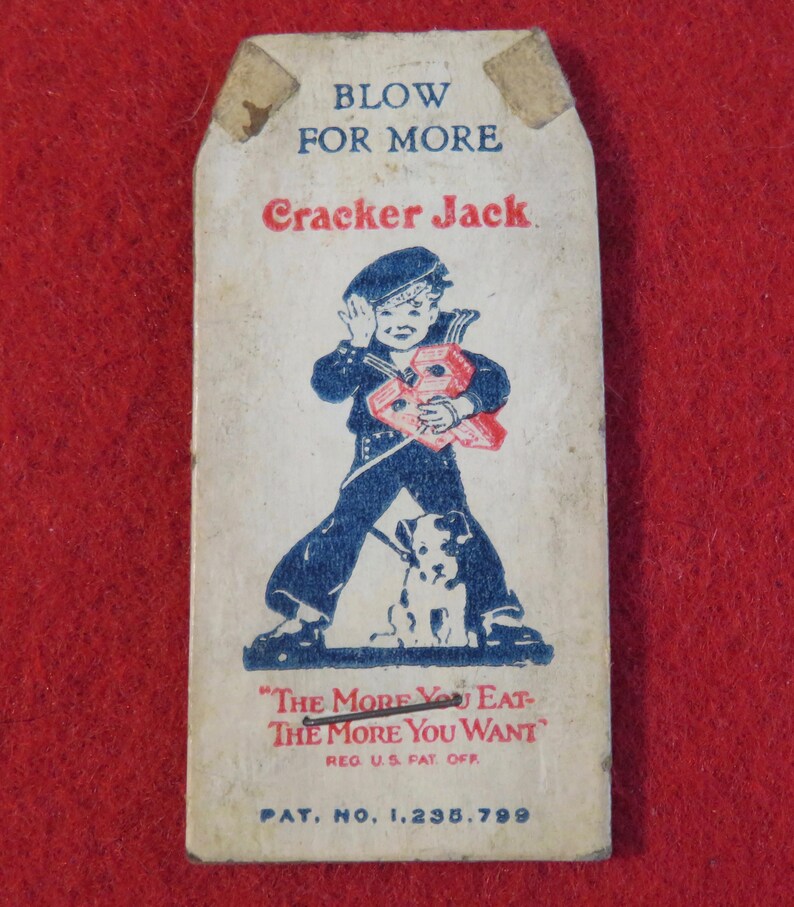 Może przedstawiać: Winietkowe pudełko Cracker Jack z niebiesko-białą ilustracją chłopca w marynarskim stroju trzymającego czerwone pudełko i psa. Na pudełku widnieje tekst "Blow for More Cracker Jack" oraz "The More You Eat-The More You Want" wraz z tekstem "Reg U.S. Pat. Off" i "Pat. No. 1,235,799".