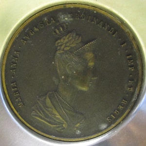 May include: A bronze-colored antique coin featuring a profile of a woman wearing a crown and a necklace. The coin is inscribed with the words "MARIA ANNA AVGSTA FERDINANDI I IMP ET REGIS" around the edge. The coin is set in a silver-colored frame.