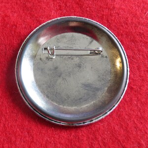 May include: The back of a silver button with a pin attached. The button is round and has a smooth surface.