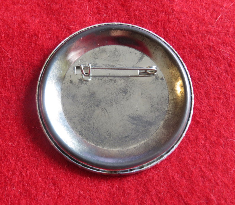 May include: The back of a silver button with a pin attached. The button is round and has a smooth surface.