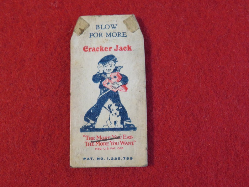 Może przedstawiać: Wierzchnia część pudełka Cracker Jack z epoki z niebiesko-białą ilustracją chłopca trzymającego czerwoną i białą nagrodę. Tekst na pudełku brzmi "Blow for More Cracker Jack" i "The More You Eat The More You Want". Numer patentu 1,235,799 jest r&oacute;wnież nadrukowany na pudełku.