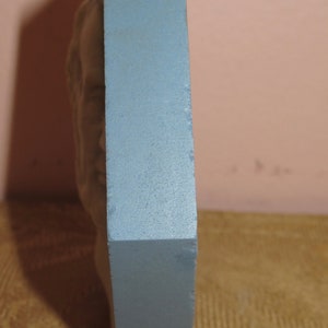 May include: A blue rectangular plaque with a raised, sculpted design of a face. The plaque is standing upright on a brown surface.