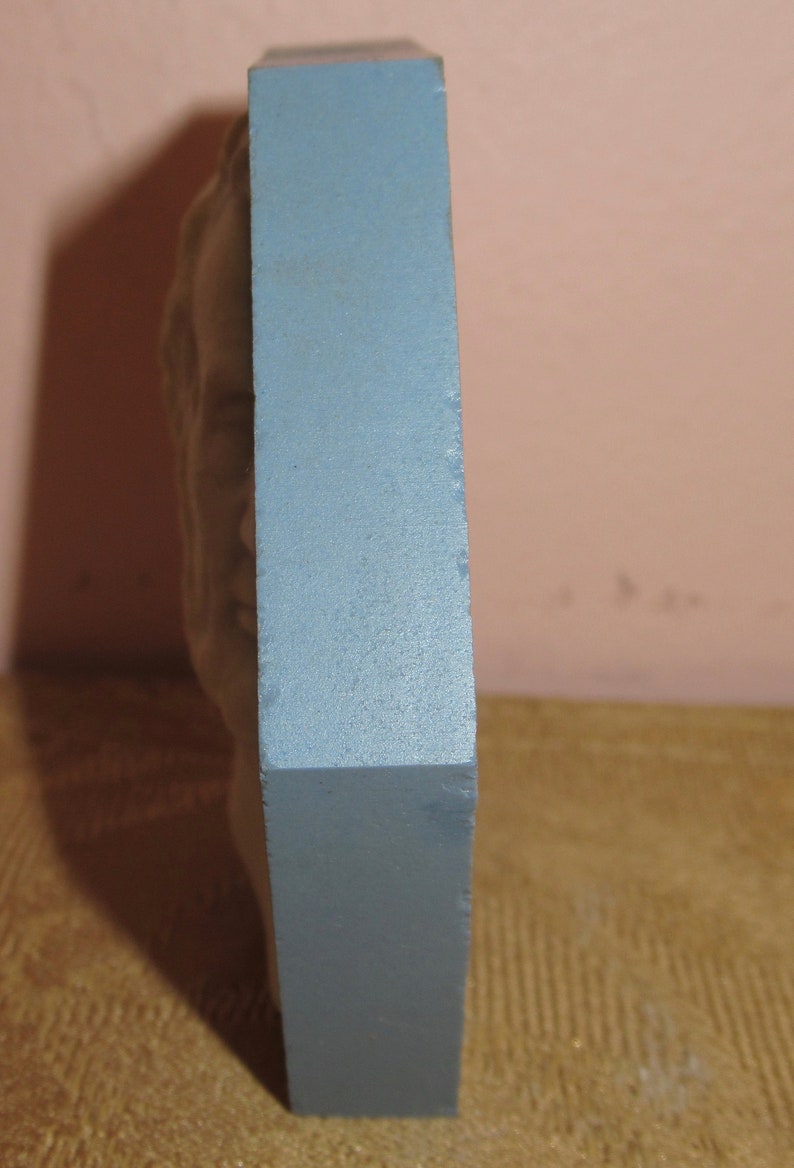 May include: A blue rectangular plaque with a raised, sculpted design of a face. The plaque is standing upright on a brown surface.