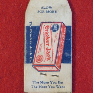 Może przedstawiać: Winietkowe pudełko Cracker Jack z tekstem "Blow for More" i "The More You Eat The More You Want". Pudełko jest czerwone, białe i niebieskie z obrazkiem orzecha.