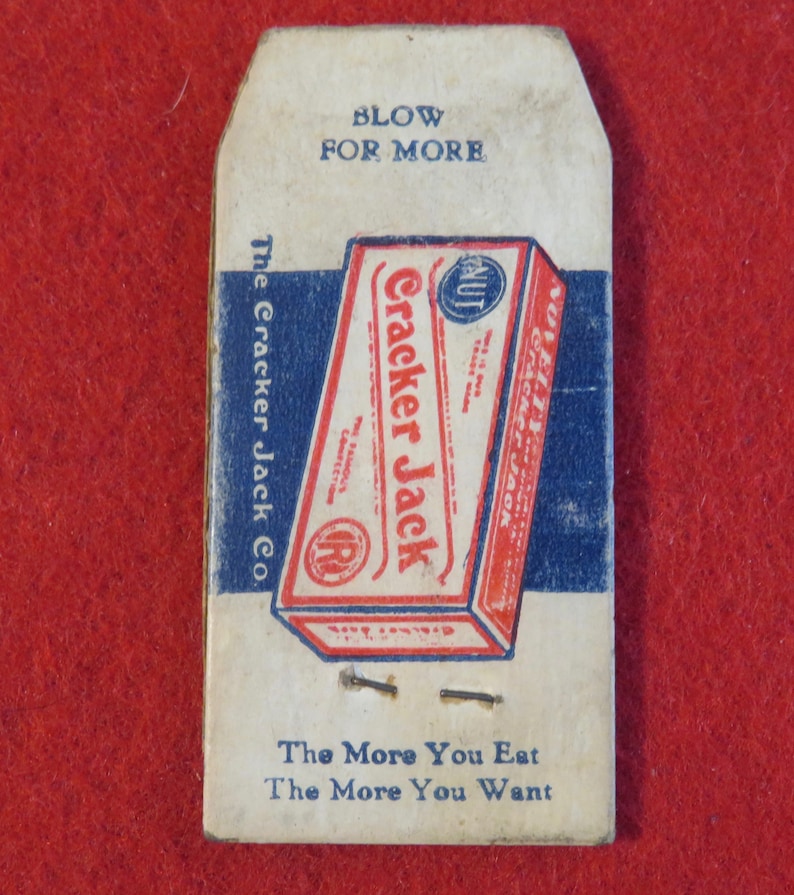 Może przedstawiać: Winietkowe pudełko Cracker Jack z tekstem "Blow for More" i "The More You Eat The More You Want". Pudełko jest czerwone, białe i niebieskie z obrazkiem orzecha.