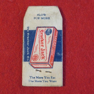 Może przedstawiać: Winietkowe pudełko Cracker Jack z tekstem "Blow for More" i "The More You Eat The More You Want". Pudełko jest czerwone, białe i niebieskie z obrazkiem pudełka Cracker Jack.