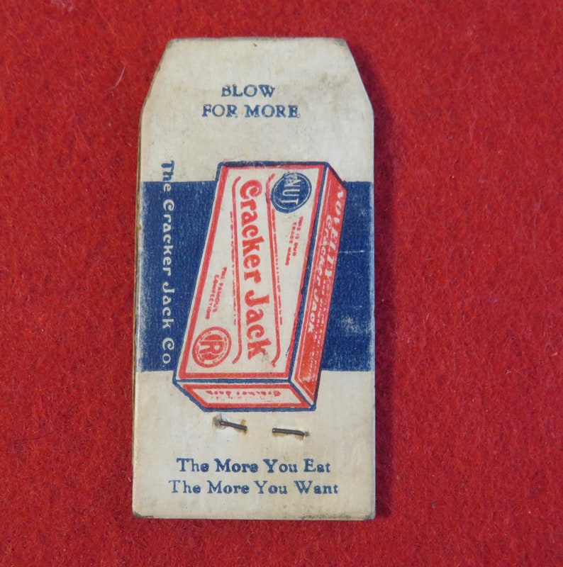 Może przedstawiać: Winietkowe pudełko Cracker Jack z tekstem "Blow for More" i "The More You Eat The More You Want". Pudełko jest czerwone, białe i niebieskie z obrazkiem pudełka Cracker Jack.