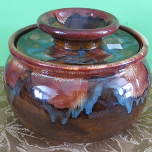 May include: A brown ceramic crock with a lid. The crock has a mottled glaze with brown, blue, and white accents. The lid has a small knob on top.