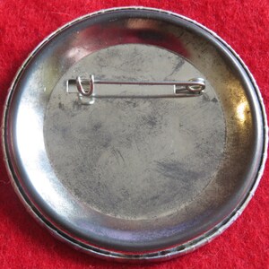 May include: The back of a silver button with a pin attached. The button has a textured rim.