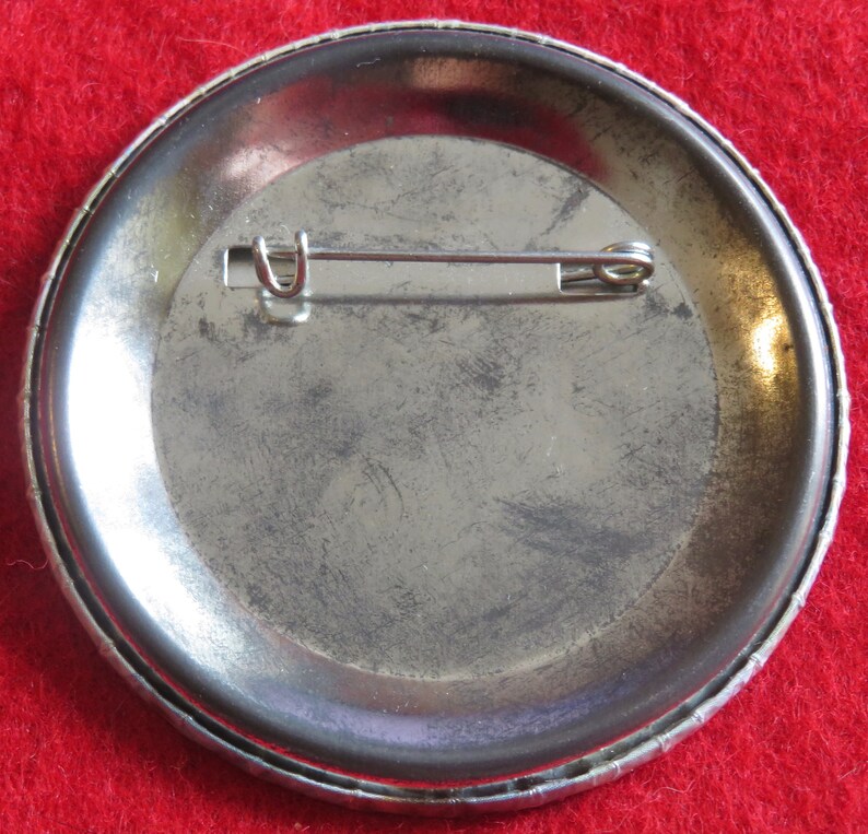 May include: The back of a silver button with a pin attached. The button has a textured rim.