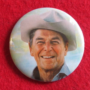 May include: A close-up of a button featuring a smiling man wearing a light gray cowboy hat. The background is a light blue sky with clouds.