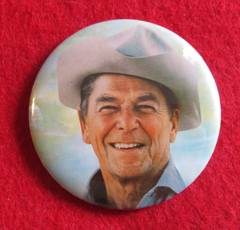 May include: A close-up of a button featuring a smiling man wearing a light gray cowboy hat. The background is a light blue sky with clouds.