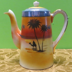 May include: A ceramic teapot with a desert scene painted on it. The scene features palm trees, a camel, and a person riding the camel. The teapot has a gold trim around the rim and handle.