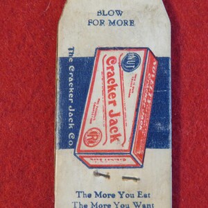 Może przedstawiać: Winietkowe pudełko Cracker Jack z niebiesko-czerwonym wzorem. Pudełko zawiera napis "Cracker Jack" czerwonymi literami i "The Famous Confection" mniejszymi białymi literami. Pudełko zawiera r&oacute;wnież niebiesko-białe k&oacute;łko z napisem "NUT" białymi literami. Pudełko znajduje się na białej kartce z napisem "Blow for More" i "The Cracker Jack Co." niebieskimi literami. Kartka zawiera r&oacute;wnież napis "The More You Eat The More You Want" niebieskimi literami.