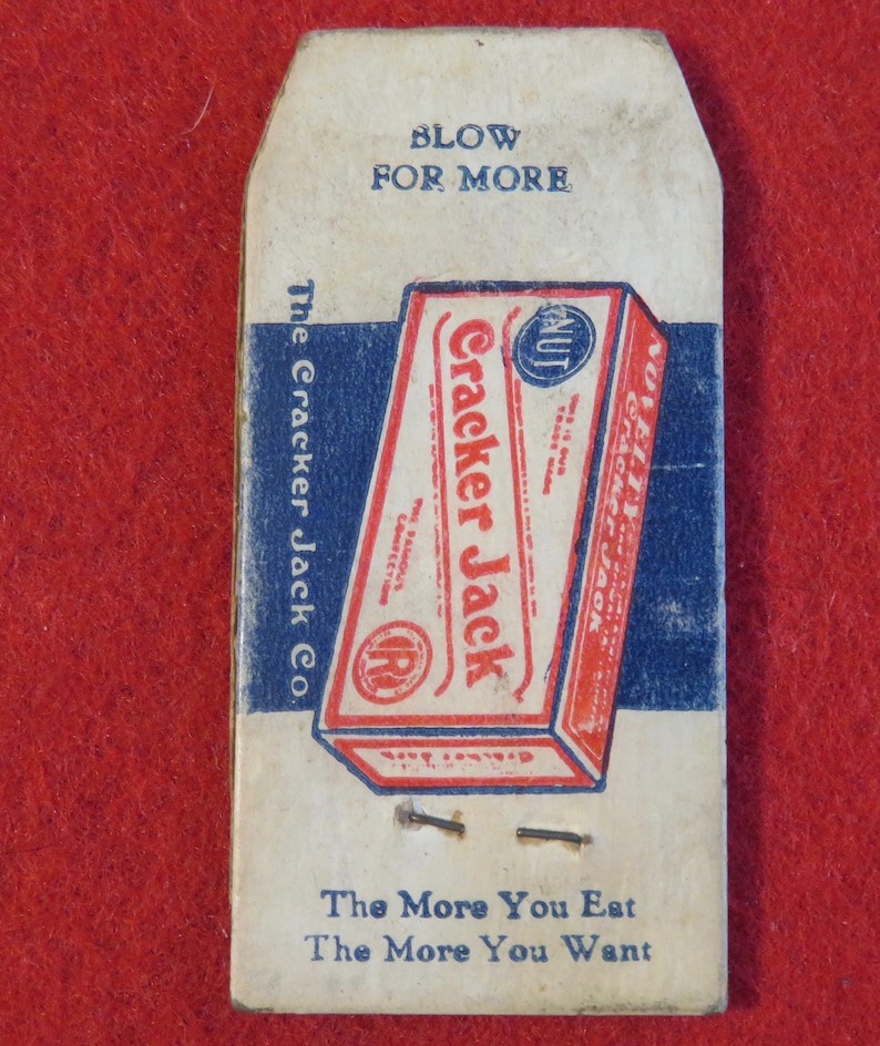 Może przedstawiać: Winietkowe pudełko Cracker Jack z niebiesko-czerwonym wzorem. Pudełko zawiera napis "Cracker Jack" czerwonymi literami i "The Famous Confection" mniejszymi białymi literami. Pudełko zawiera r&oacute;wnież niebiesko-białe k&oacute;łko z napisem "NUT" białymi literami. Pudełko znajduje się na białej kartce z napisem "Blow for More" i "The Cracker Jack Co." niebieskimi literami. Kartka zawiera r&oacute;wnież napis "The More You Eat The More You Want" niebieskimi literami.