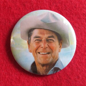 May include: A close-up of a button featuring a smiling man wearing a cowboy hat. The background is a light blue sky with clouds.