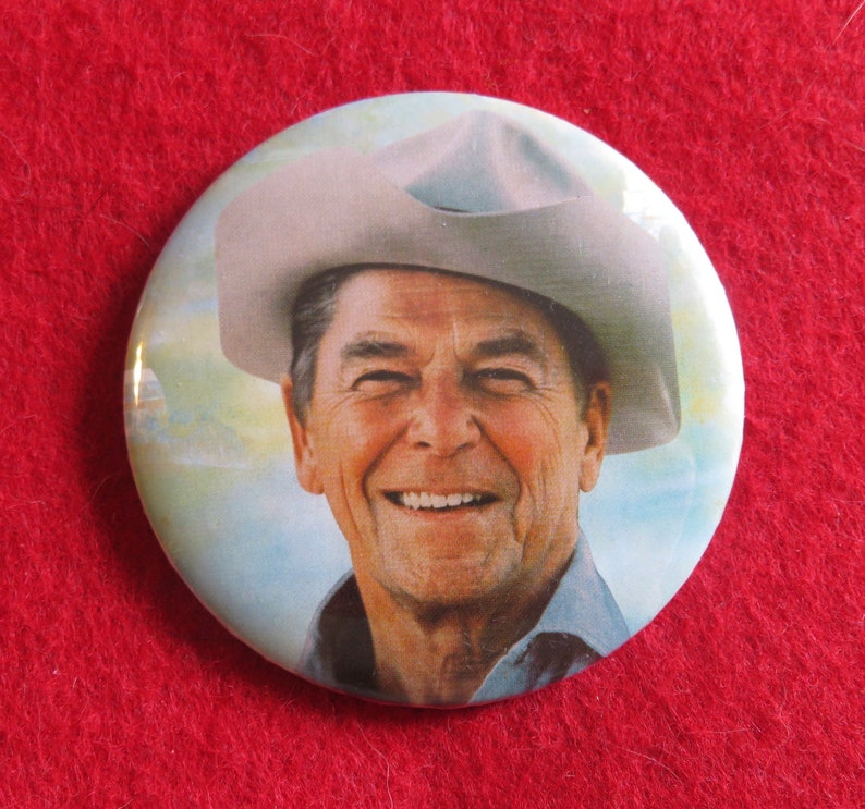 May include: A close-up of a button featuring a smiling man wearing a cowboy hat. The background is a light blue sky with clouds.
