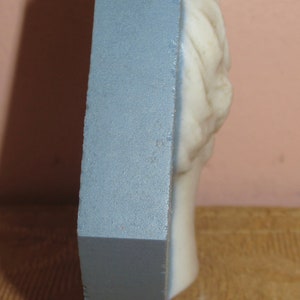 May include: A white plaster bust of a person with a detailed hairstyle. The bust is mounted on a blue rectangular base.