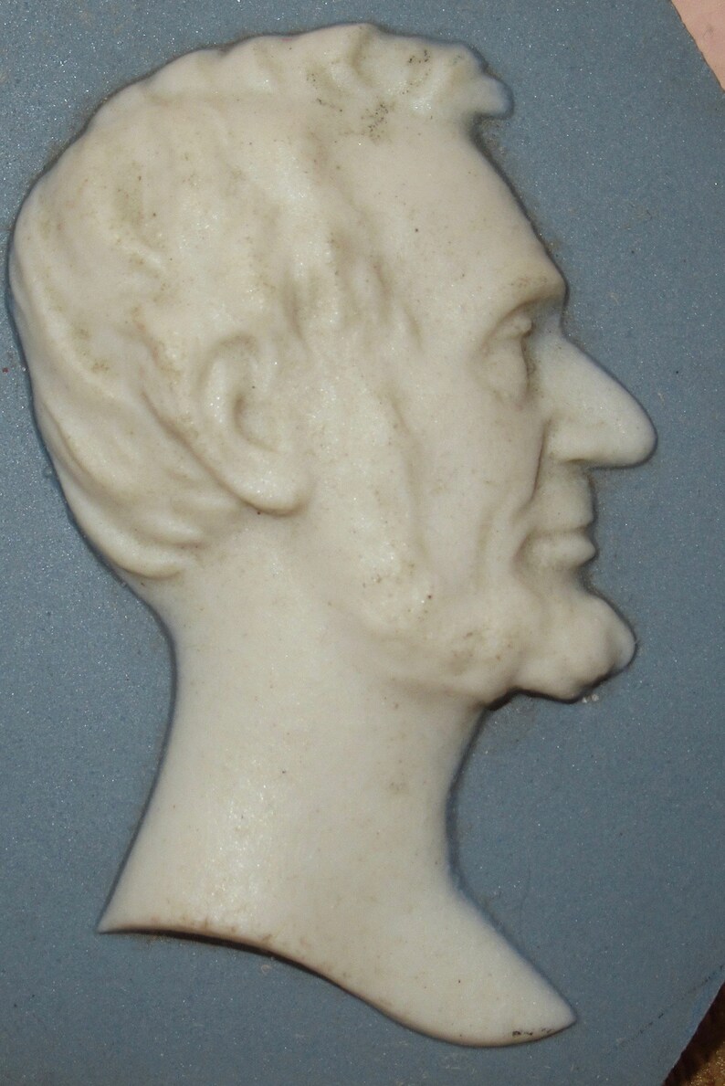 May include: A white porcelain cameo of a man's profile, with a detailed face and hair, set against a blue background.