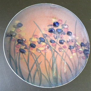 May include: A round ceramic plate with a floral design. The plate features a watercolor-like painting of blue, pink, and yellow flowers with green stems on a peach background.