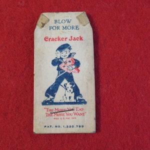 Może przedstawiać: Wierzchnia część pudełka Cracker Jack z epoki z chłopcem w marynarskim stroju trzymającym pudełko Cracker Jack i psa. Tekst na pudełku brzmi "Blow for More Cracker Jack. "The More You Eat The More You Want." Reg. U.S. Pat. Off. Pat. No. 1,235,799."