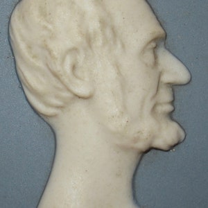 May include: A white ceramic cameo of a man's profile, with a detailed face and beard, set against a blue background.