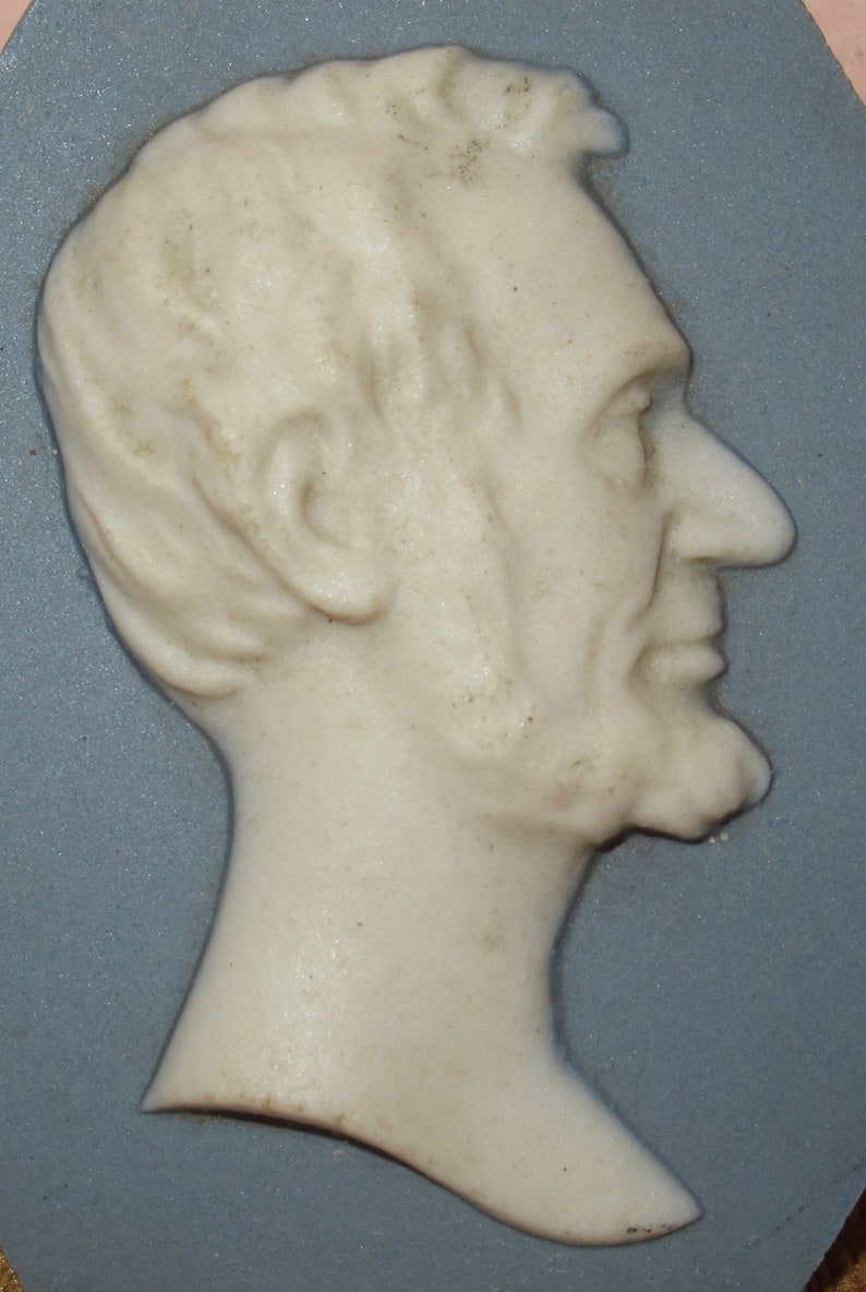 May include: A white ceramic cameo of a man's profile, with a detailed face and beard, set against a blue background.