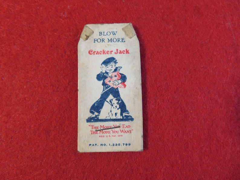 Może przedstawiać: Wierzchnia część pudełka Cracker Jack z epoki z niebiesko-białą ilustracją marynarza trzymającego worek Cracker Jack i psa. Tekst na g&oacute;rze pudełka brzmi "Blow for More Cracker Jack" i "The More You Eat The More You Want". Numer patentu jest r&oacute;wnież uwzględniony: "Pat. No. 1,235,799".