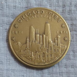 May include: A gold-colored commemorative coin with the text "CHICAGO FIRE CENTENNIAL" and the years 1871 and 1971. The coin features a detailed depiction of the Chicago skyline and is from the Chicago Historical Society.
