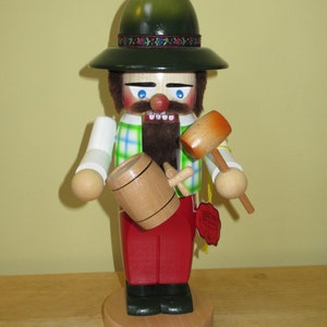 May include: A wooden nutcracker figurine with a green hat, red and white plaid shirt, and a red beard. The nutcracker is holding a wooden mallet and a barrel.