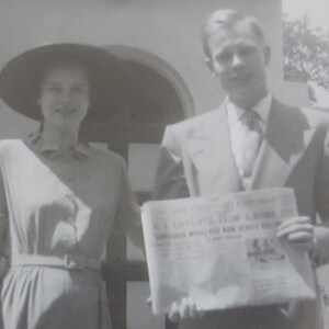 May include: Black and white photograph of a couple posing outdoors. The woman wears a dress and a wide-brimmed hat. The man holds a newspaper with visible text. The image has a vintage aesthetic.