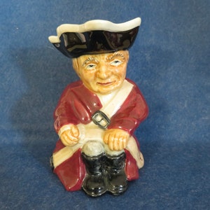 May include: A ceramic figurine of a man wearing a black and white tri-cornered hat, a red jacket with white trim, and black boots. He is sitting with his legs crossed and holding a white object in his hand.