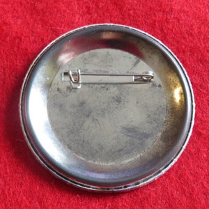 May include: The back of a silver button with a pin attached. The button has a smooth, slightly textured surface.