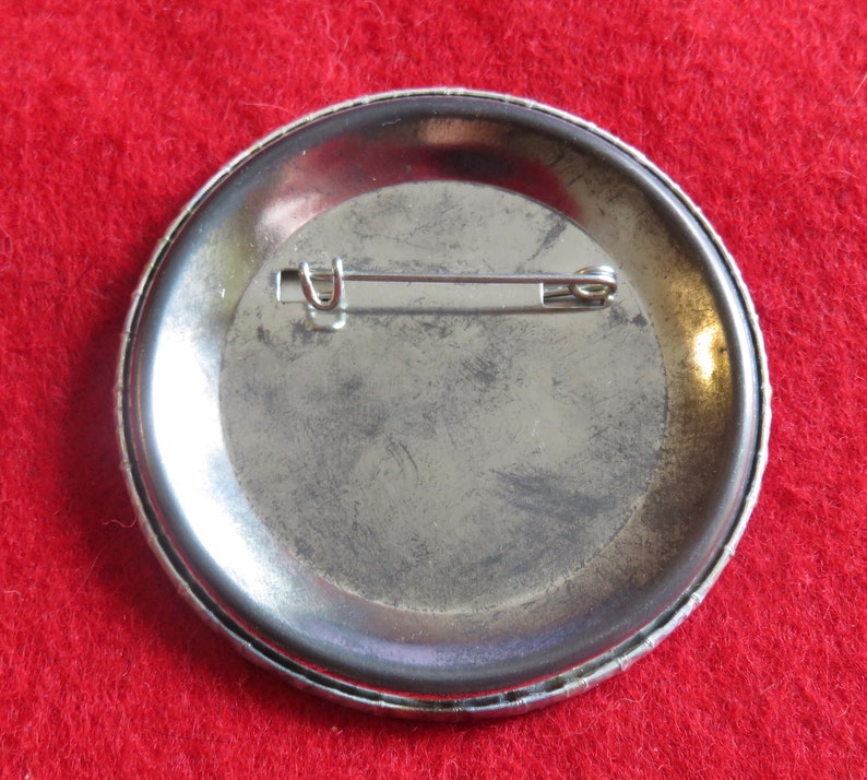 May include: The back of a silver button with a pin attached. The button has a smooth, slightly textured surface.