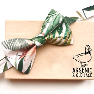 Può includere: Un papillon bianco con un motivo a foglie tropicali verdi e arancioni. Il papillon è annodato e mostrato su una scatola marrone con il testo "ARSENIC & OLD LACE" e un'illustrazione in bianco e nero di un'anatra con papillon.