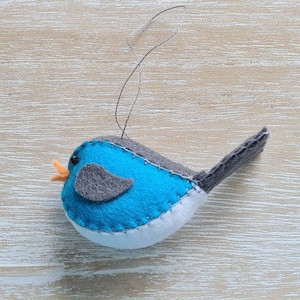 May include: A blue felt bird ornament with a grey wing and orange beak. The bird has a white felt belly and a loop for hanging.