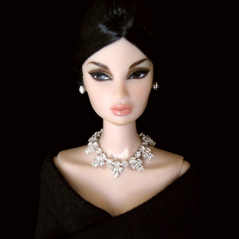 May include: A doll with black hair and dark makeup is wearing a black dress and a sparkling silver necklace.