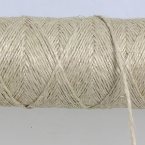 May include: A spool of natural beige linen thread. The thread is wound tightly around the spool and a long strand of thread hangs off the end.