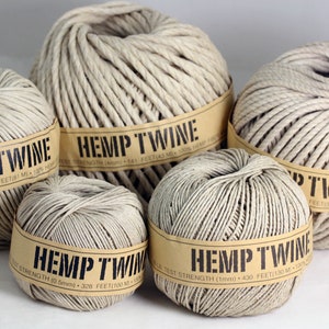 Puede incluir: Cuatro carretes de cuerda de cáñamo natural de diferentes tamaños. Cada carrete tiene una etiqueta de papel marrón con el texto "Hemp Twine" y detalles sobre la resistencia y la longitud de la cuerda en metros.