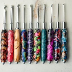 May include: A collection of ten crochet hooks arranged in a row, each featuring a metallic silver hook and a unique, contoured handle. The handles display various patterns and colors, including marbled resin, natural wood grain, and floral designs.