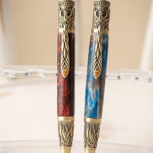 May include: Two ornate pens with intricate metalwork and a phoenix design. One pen has a red and brown wood grain pattern, while the other has a blue and white wood grain pattern.