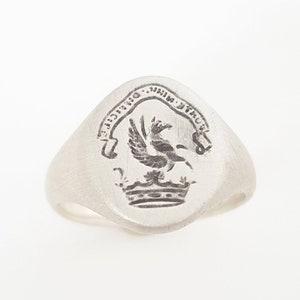 May include: A silver signet ring with an oval shaped top featuring an engraved crest with a bird and a crown. The crest is surrounded by a banner with the text "MONT.  DRACUT.  1710."