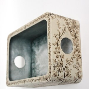 May include: A rectangular off-white wall-mounted box featuring a dark brown ink illustration of climbing ivy vines on its exterior. The interior is painted in a muted grey-blue tone. The brand name Krisoft is printed on the side of the box.