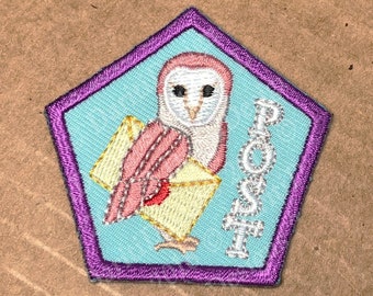 Puede incluir: Parche bordado en forma de pentágono con borde morado. El parche representa un búho rosa y blanco sosteniendo un sobre amarillo. La palabra "POST" está bordada en plata. El texto "WoRM Scouts Badge/Patches ©DaritiDesigns" está arriba y abajo.