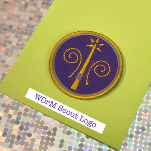 May include: A circular embroidered patch with a purple background and a gold wand design. The patch has a gold border and the text "WoRM Scout Logo" in a white rectangle.