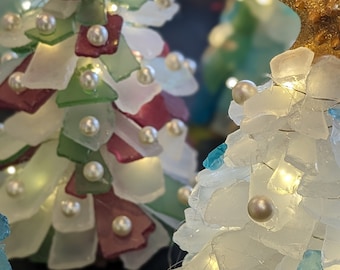 May include: Three small, decorative Christmas trees made of sea glass and white pearls. The trees are lit with white fairy lights and topped with gold starfish.
