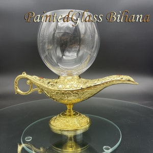 May include: A gold genie lamp with a clear glass globe on top. The lamp has intricate details and a red jewel on the spout. The lamp is sitting on a clear glass surface.